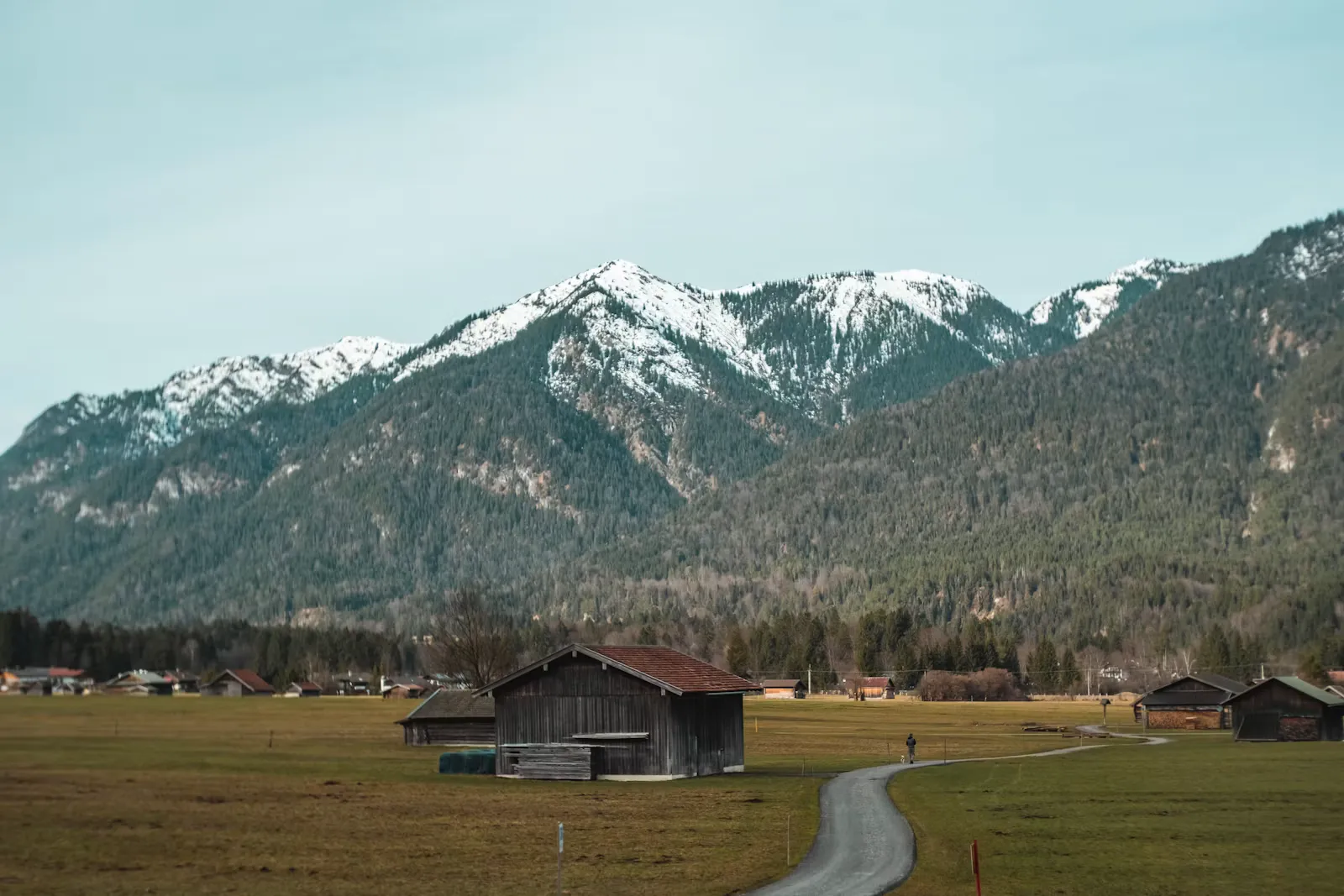 Holzstadel auf einer weiten Wiese, dahinter ein schneebedeckter Bergkamm und ein schmaler Feldweg