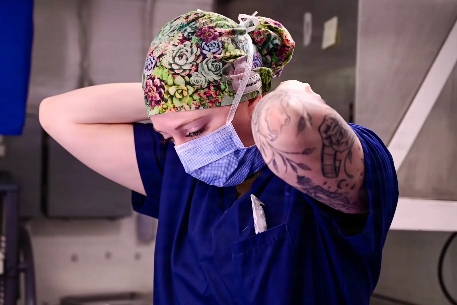 Health worker in blue scrubs and a patterned surgical cap reaching behind her head to tie a blue surgical mask