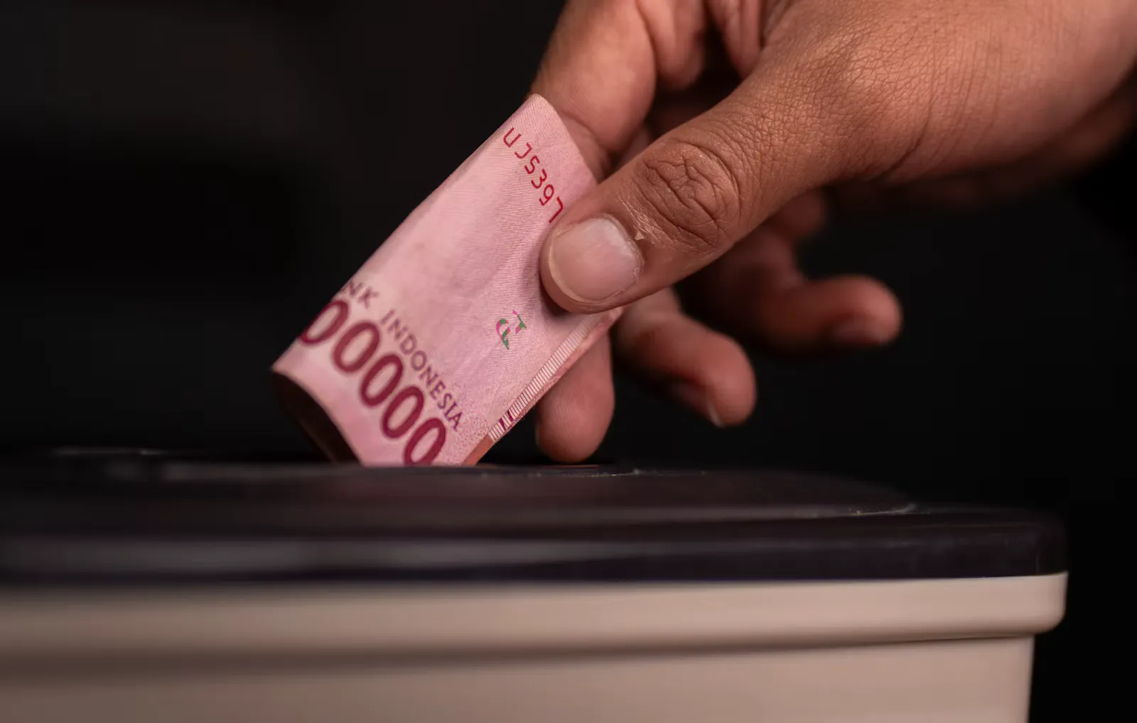 A hand pushing a folded pink 100,000 rupiah banknote from Bank Indonesia into the slot of a box
