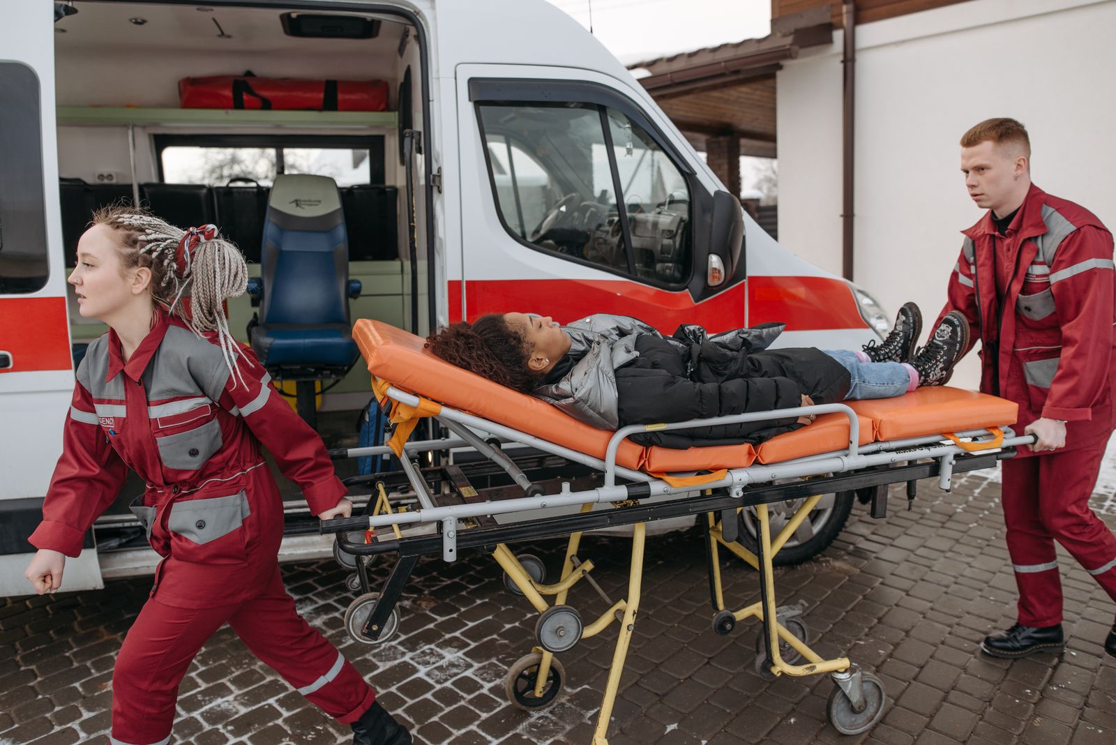 Zwei Rettungskräfte in roter Einsatzkleidung schieben eine Patientin auf einer Trage vom Rettungswagen weg