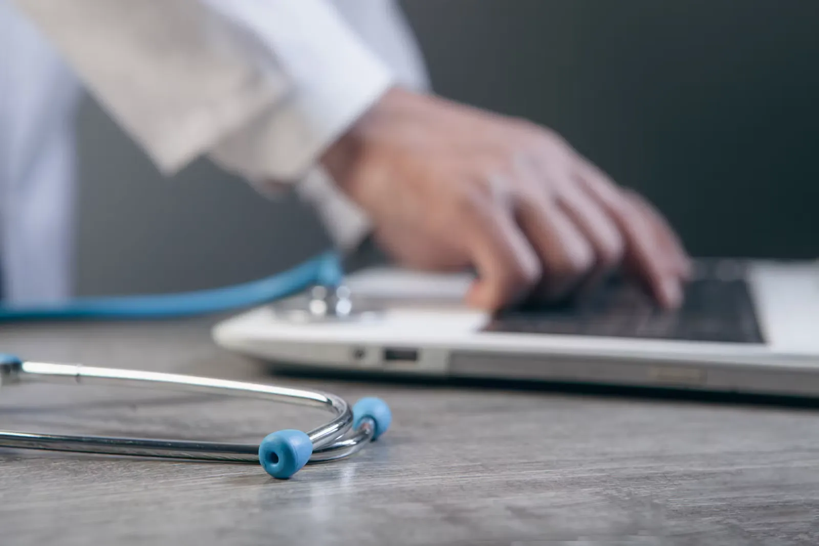 Doctor in a white coat typing on a laptop with a stethoscope lying on the desk in the foreground