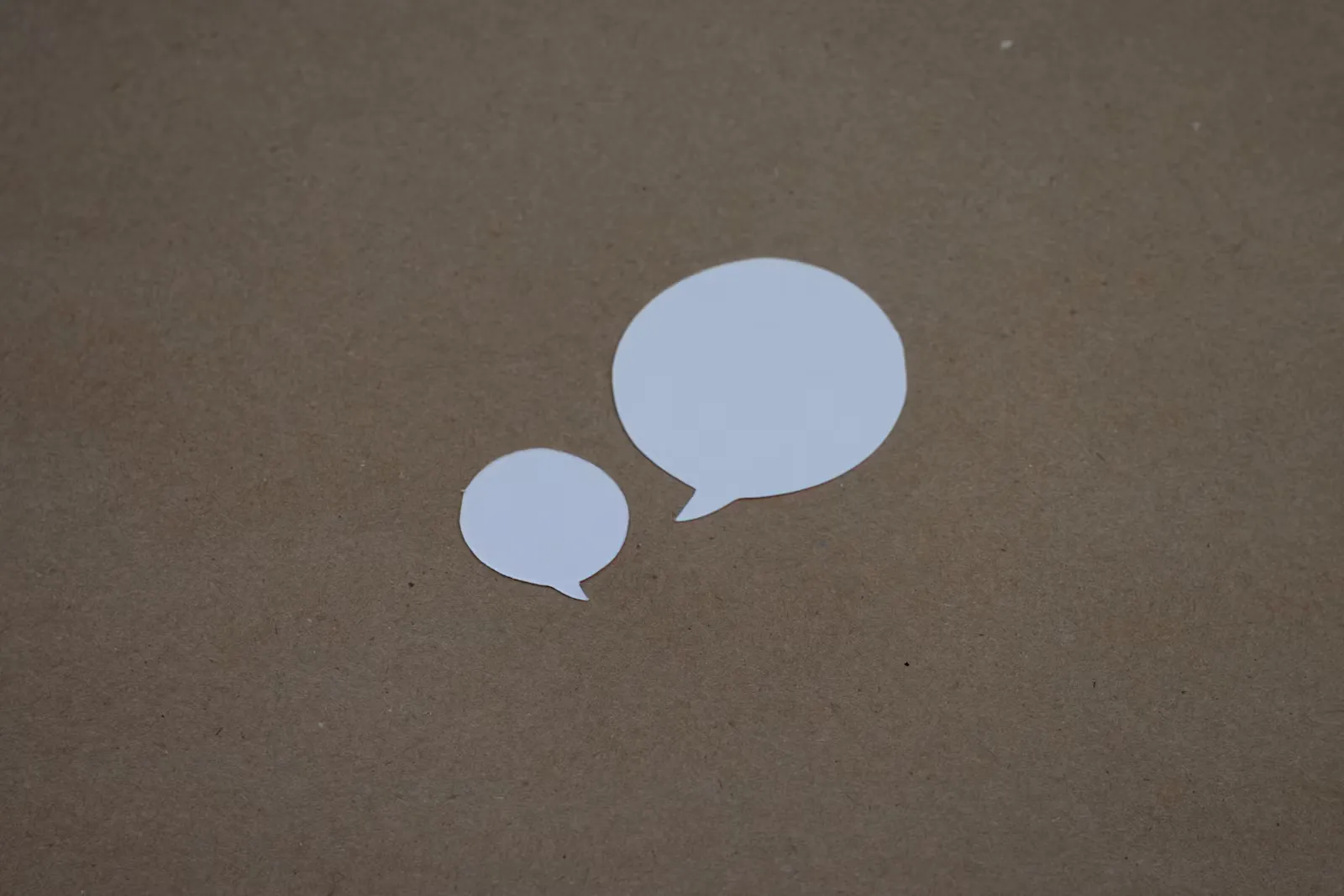 Zwei weiße Sprechblasen aus Papier, eine große und eine kleine, auf braunem Karton