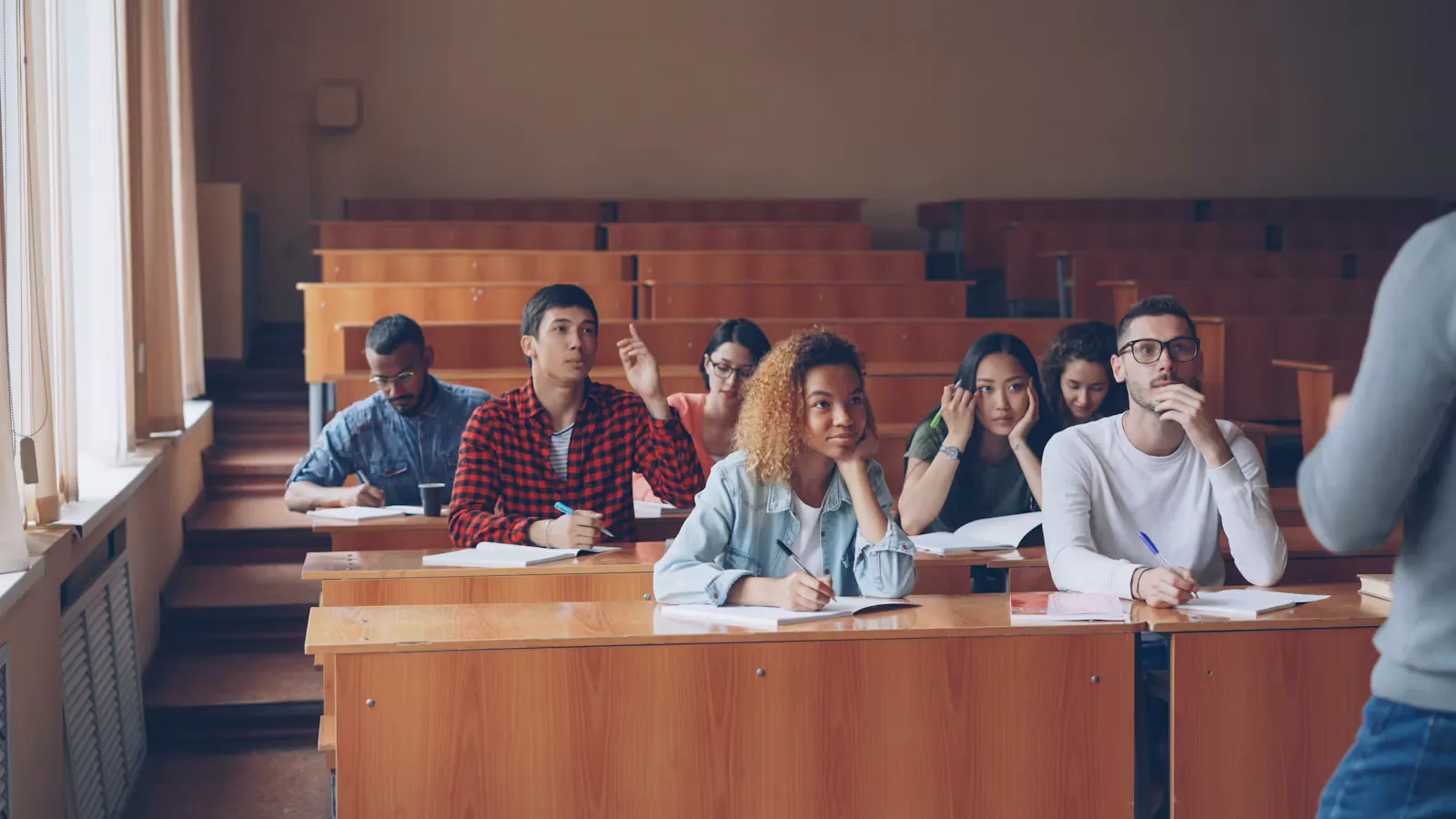 Internationale Studierende sitzen in einem Hörsaal mit Holzbänken und hören aufmerksam zu, eine Studentin meldet sich