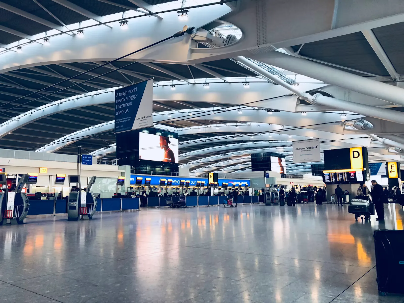 A wide airport departure area with a large departures board and passengers waiting below it