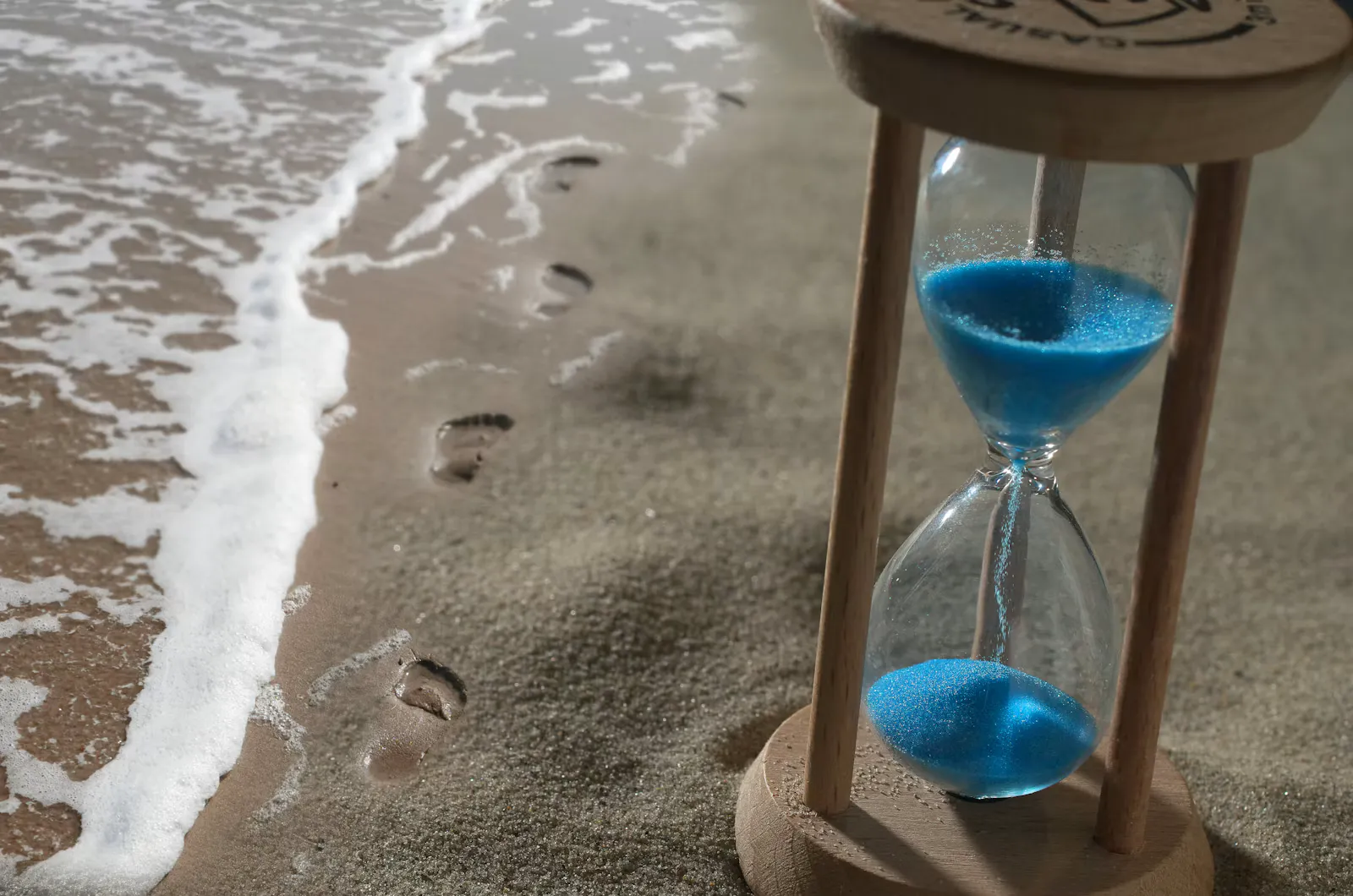 Wooden hourglass with blue sand standing on a beach next to footprints and the incoming surf