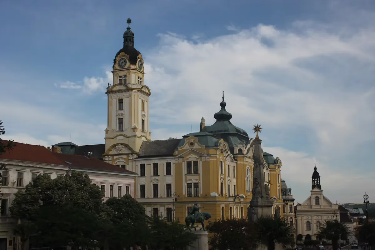 The yellow town hall of Pécs with its clock tower, a bronze equestrian statue and a stone column on Széchenyi Square under a cloudy sky