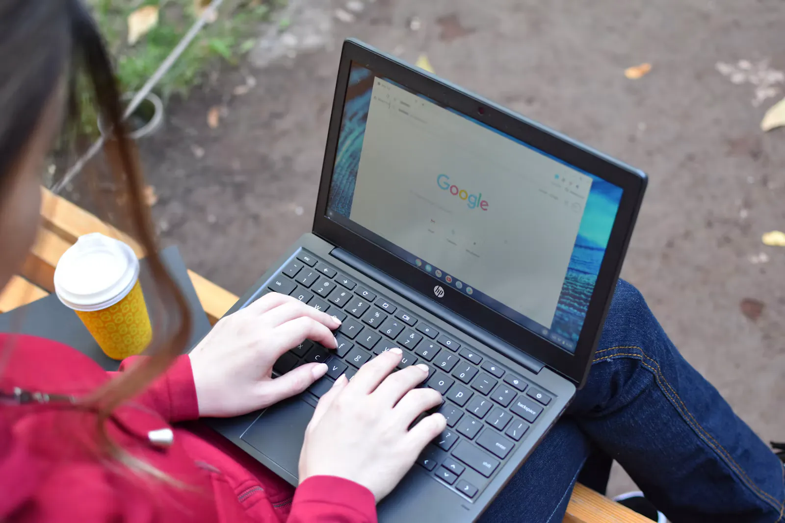 Studentin im roten Pullover tippt auf einem Laptop, der auf ihren Knien liegt, daneben steht ein gelber Kaffeebecher auf einer Holzbank im Freien