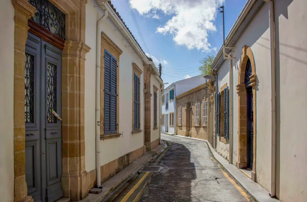 Quiet old town street in Nicosia with pale stone houses, tall grey wooden shutters, arched doorways and a narrow road under a blue sky with white clouds