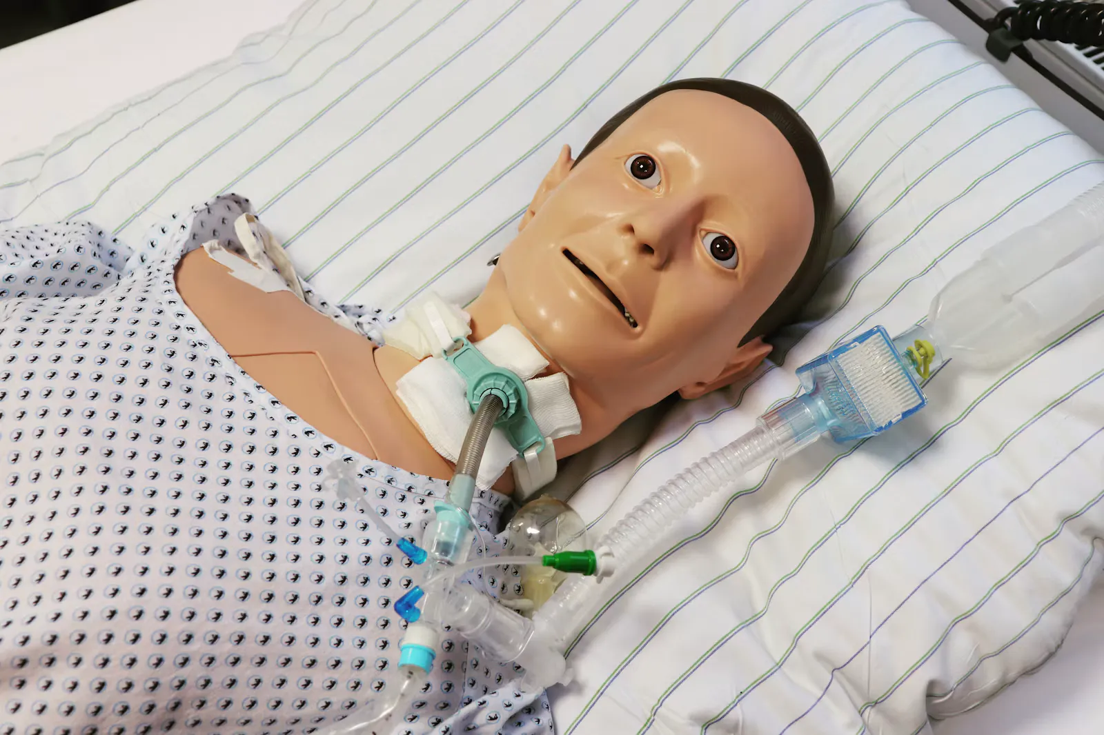 Close up of a patient simulator manikin with a tracheostomy tube and suction tubing lying on a hospital bed with striped pillows