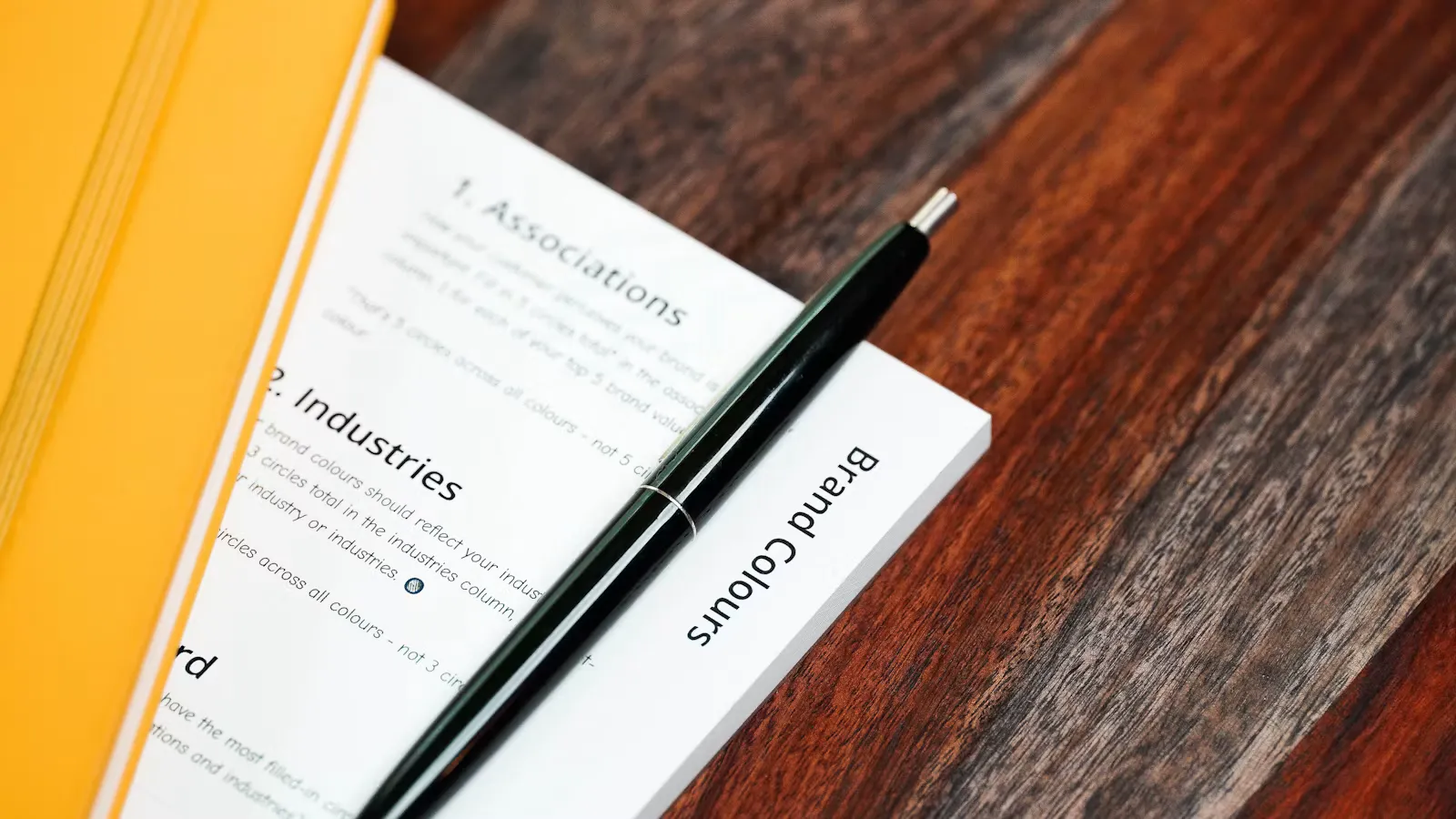 Englischsprachiges Arbeitsblatt mit schwarzem Kugelschreiber neben einem gelben Notizbuch auf einem dunklen Holztisch