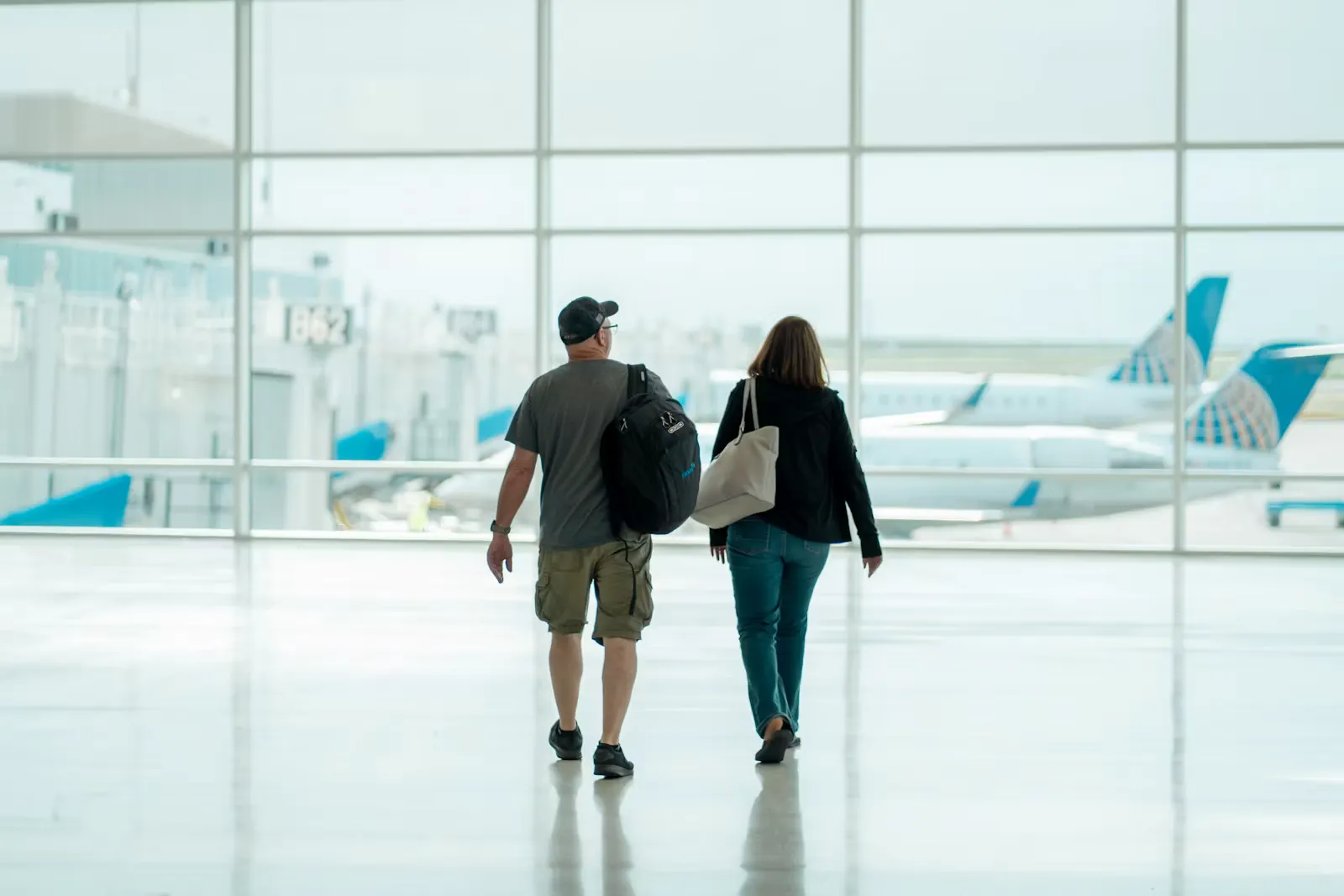 Zwei Reisende mit Rucksack und Tasche gehen durch eine helle Flughafenhalle auf die Fensterfront zu, dahinter stehen Flugzeuge am Gate