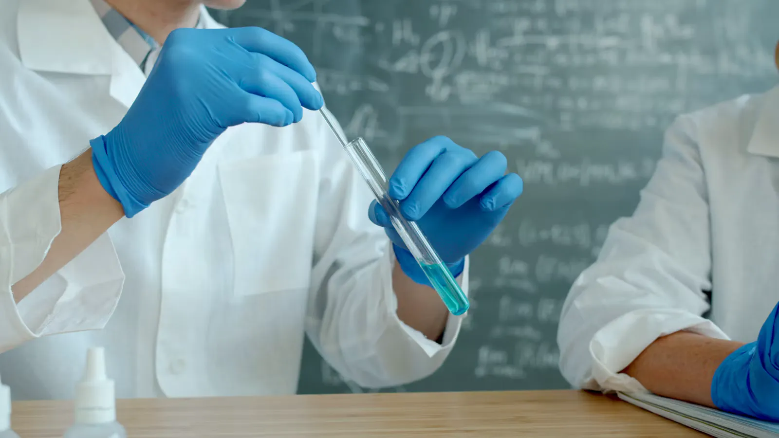 Two people in white lab coats and blue gloves, one holding a test tube of blue liquid in front of a chalkboard, a classroom view of biotechnology principles and processes