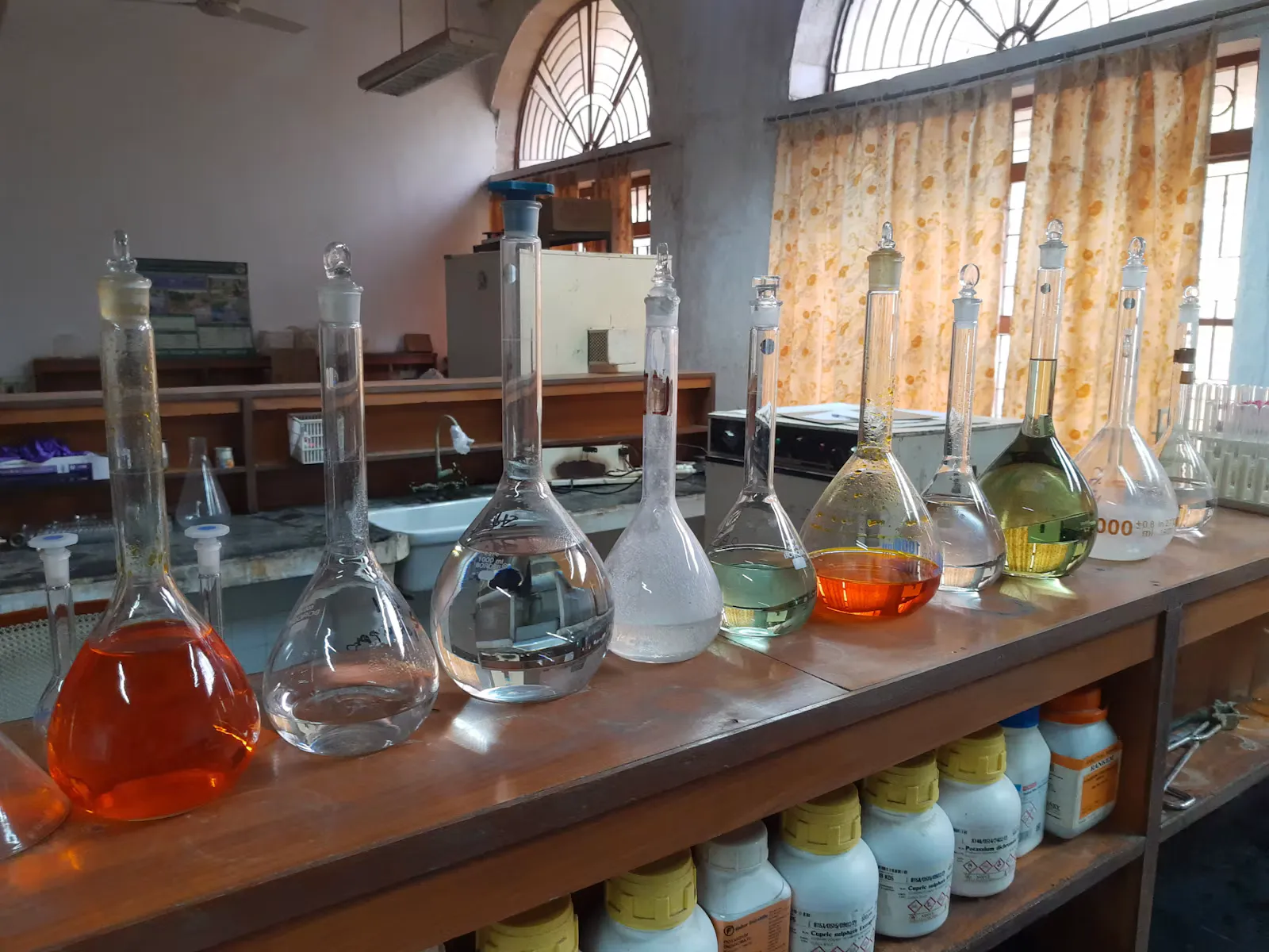 A row of volumetric flasks holding orange A row of volumetric flasks holding orange, green and clear solutions on the bench of a school chemistry lab, where the reagents behind name reactions for NEET are first met