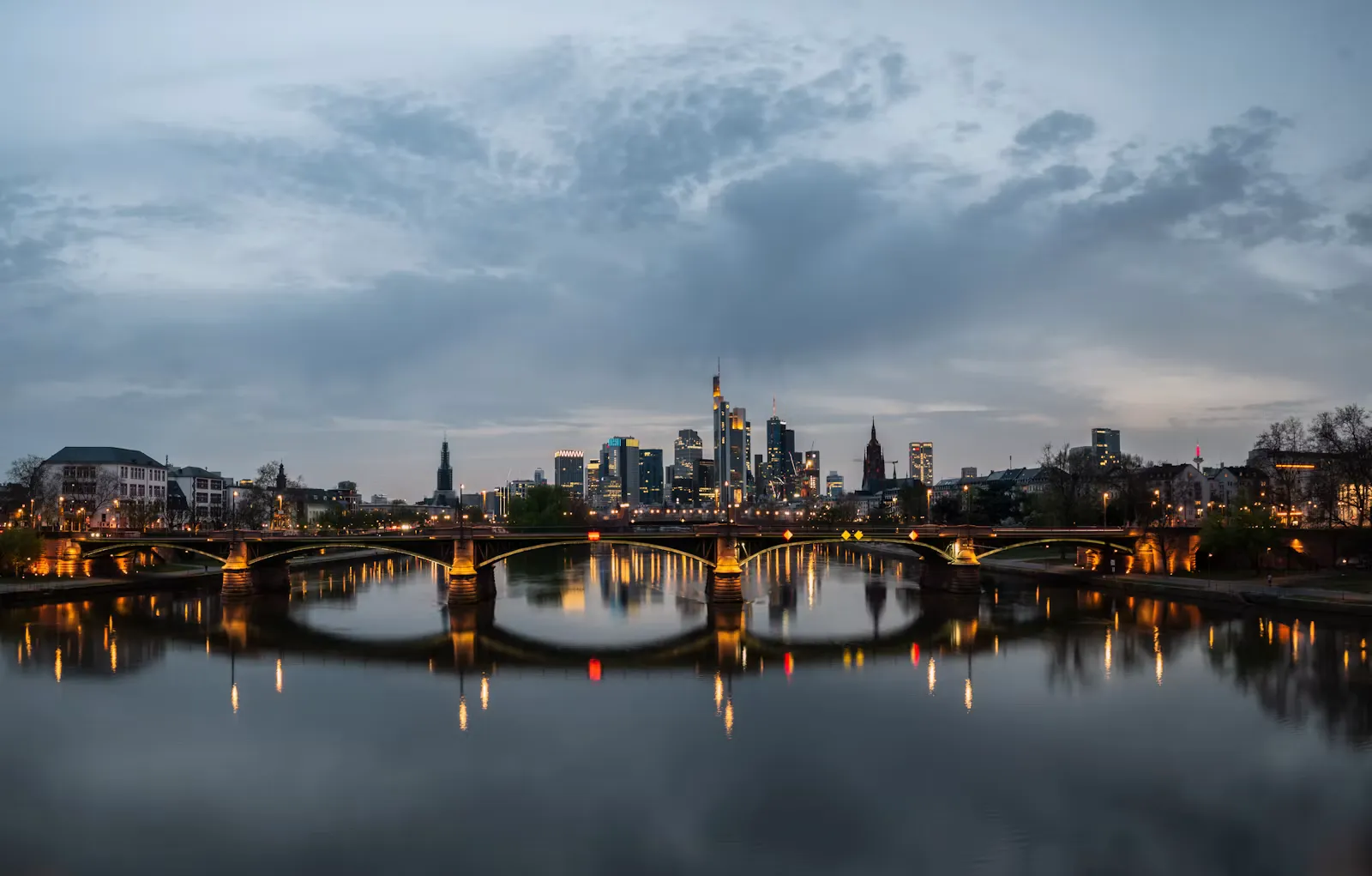 Frankfurter Skyline in der Abenddämmerung mit beleuchteter Brücke über den Main und Spiegelungen im Wasser