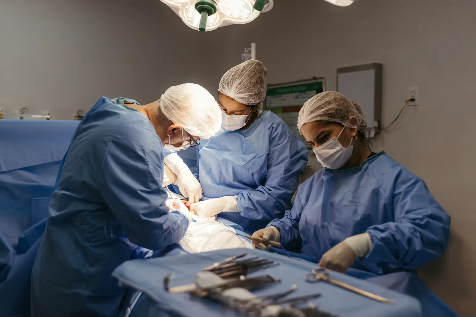 Drei Chirurginnen und Chirurgen in blauer Schutzkleidung mit Hauben und Masken operieren unter einer OP-Leuchte, vorne liegt das Instrumentenbesteck