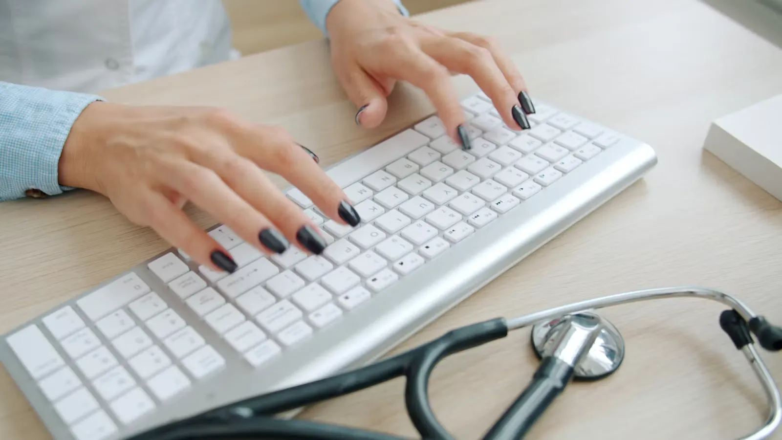 Hands typing on a white keyboard with a stethoscope lying on the desk beside it