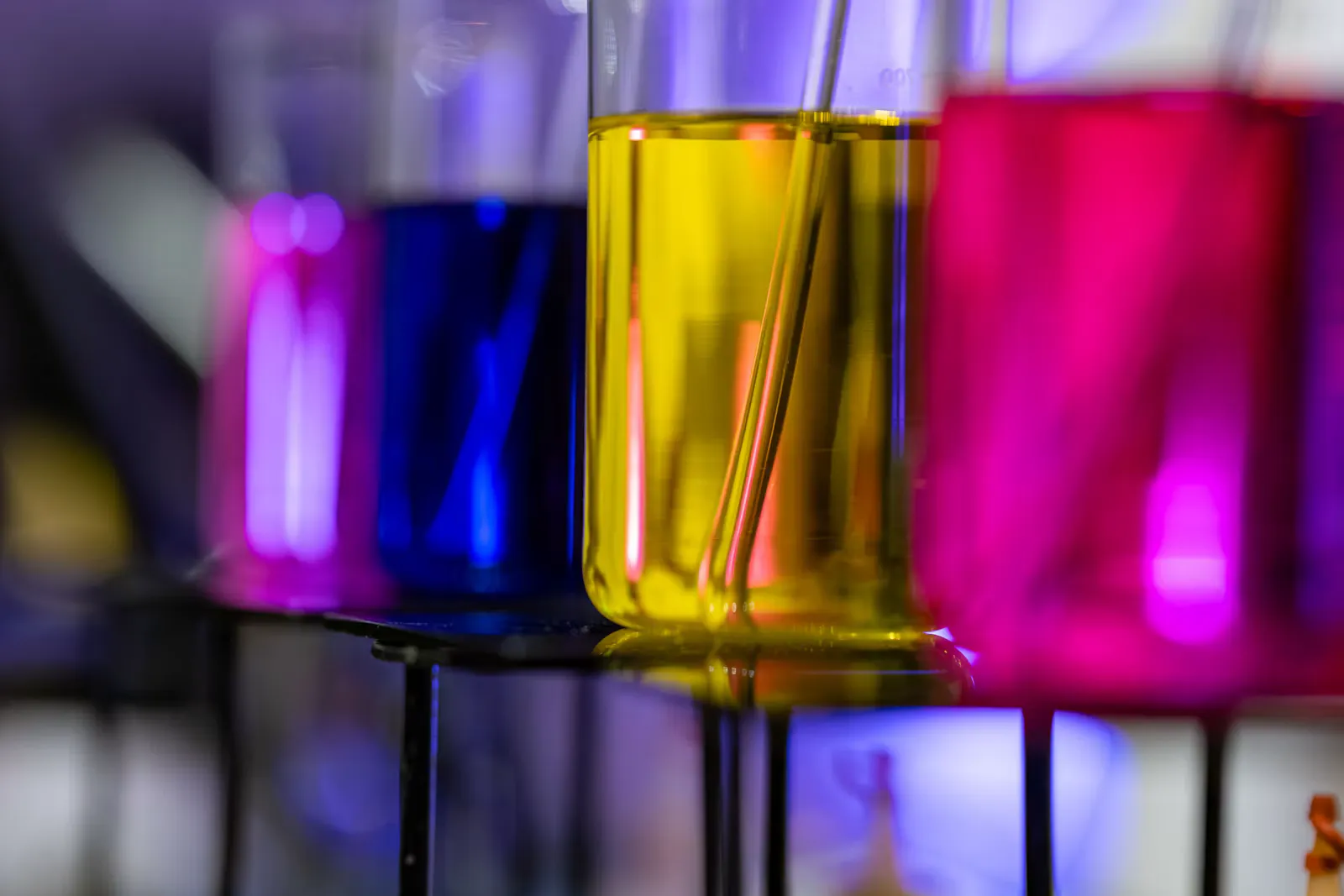 Close-up of laboratory beakers holding bright yellow, pink and blue liquids on a metal rack, a reminder that p block elements chemistry is about patterns across a row