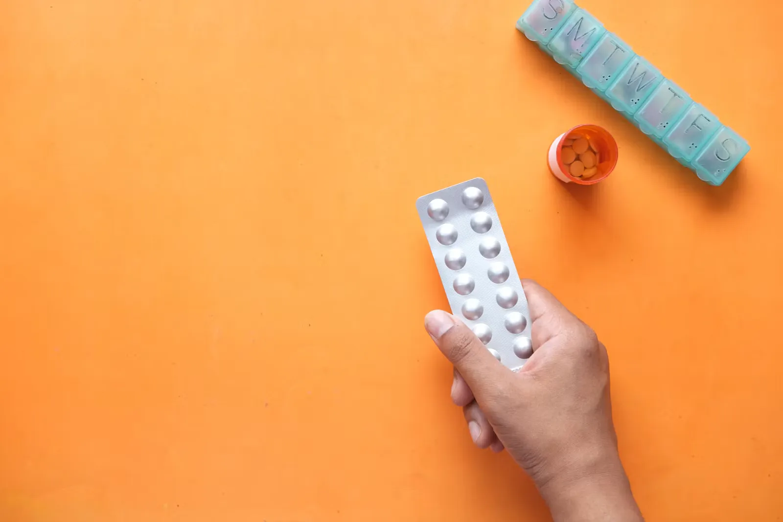 Hand hält einen silbernen Tablettenblister über einer orangen Fläche, daneben eine Wochendosierbox und ein geöffnetes Tablettenfläschchen
