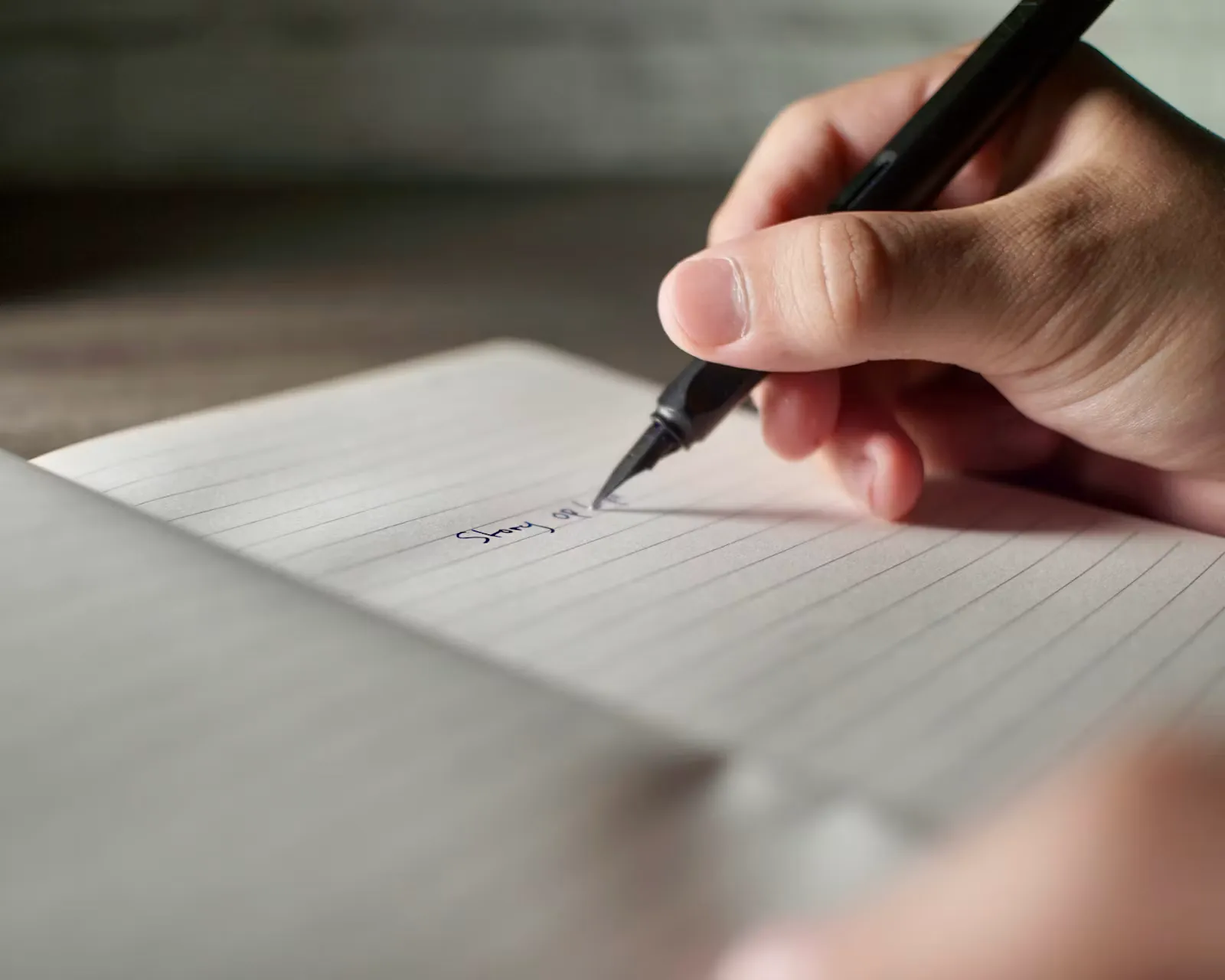 A hand holding a black pen writing on a lined page in an open notebook, the first words of a draft visible