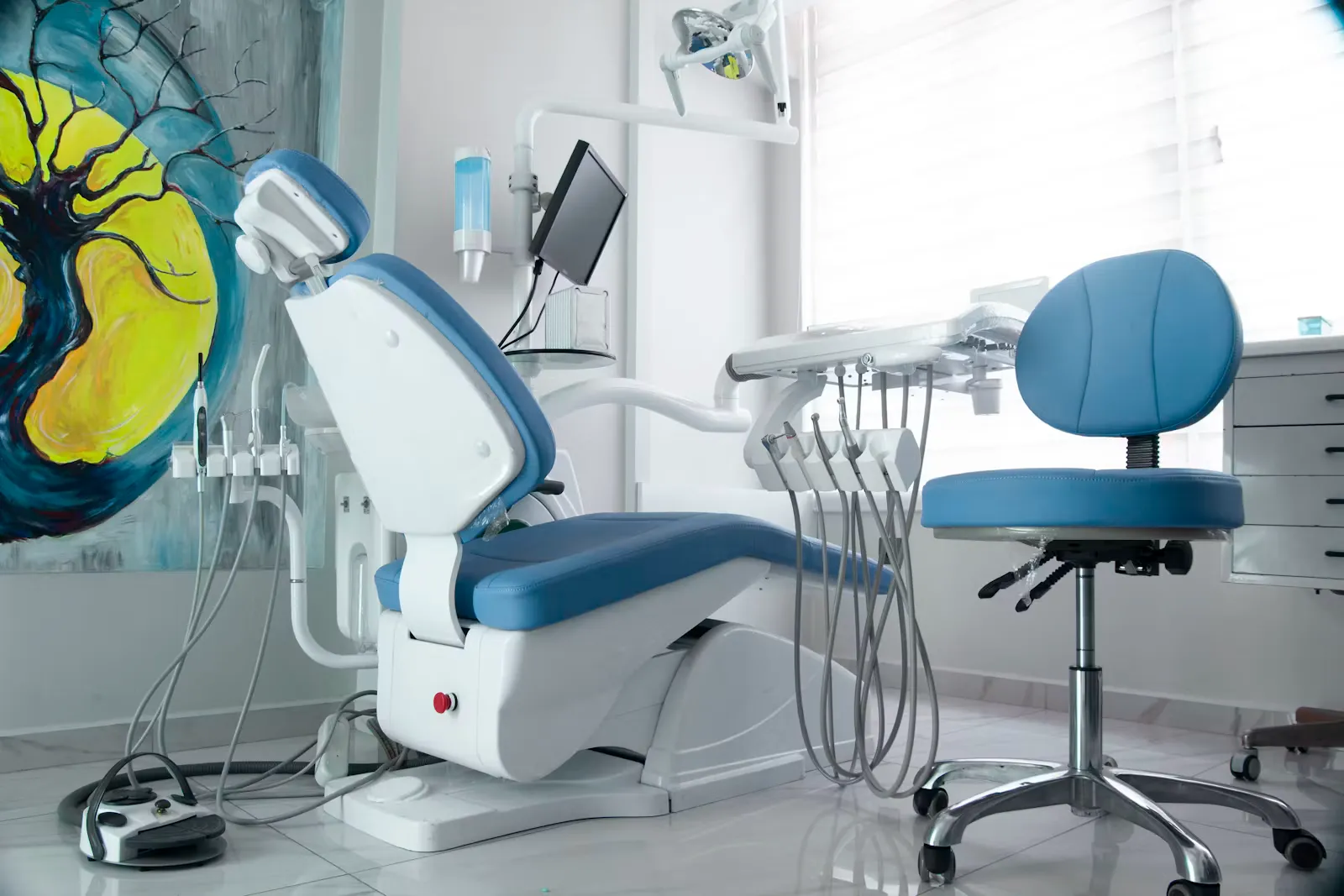 An empty dental surgery with a blue and white treatment chair, an instrument arm and hoses beside it, a dentist's stool on the right and a painted canvas on the wall