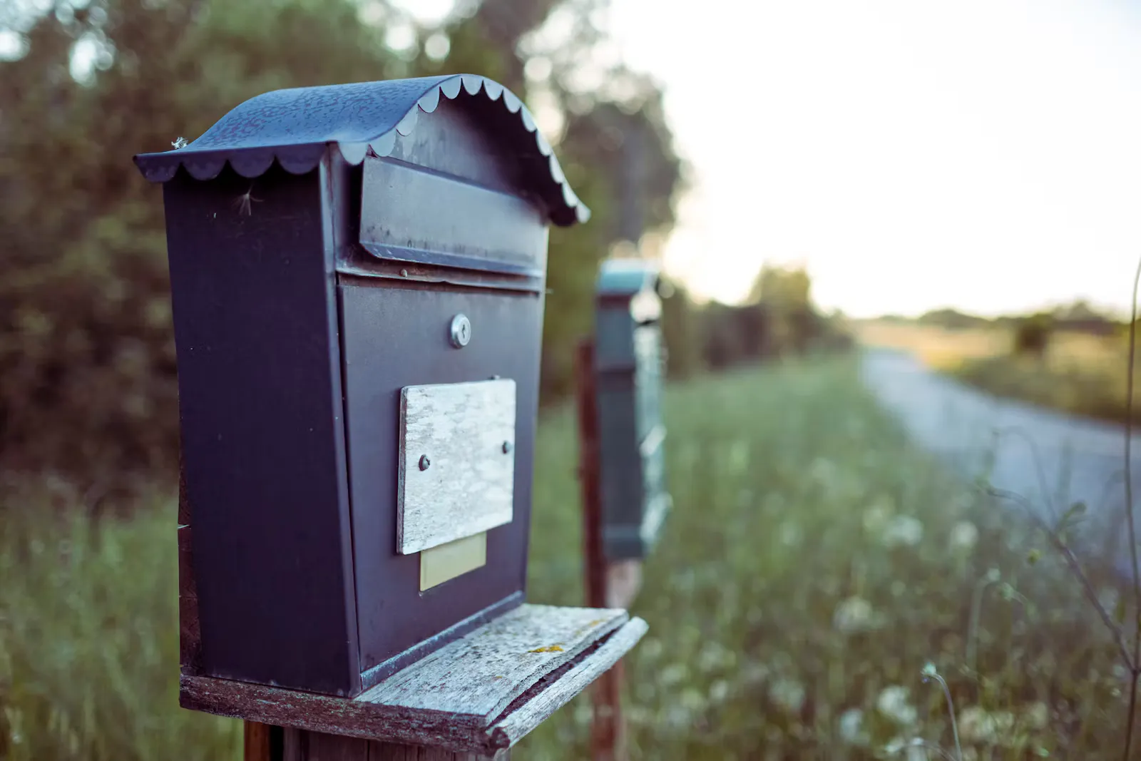 Dunkler Briefkasten aus Metall auf einem Holzpfosten am Rand eines grünen Feldwegs im Abendlicht