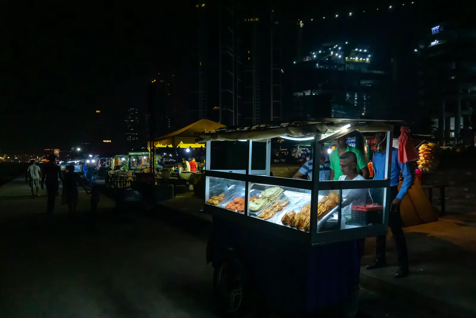 Beleuchteter Imbisswagen mit Glasvitrine an einer nächtlichen Strandpromenade in Sri Lanka, in der Vitrine liegen fertig zubereitete Fladenbrote und frittierte Snacks