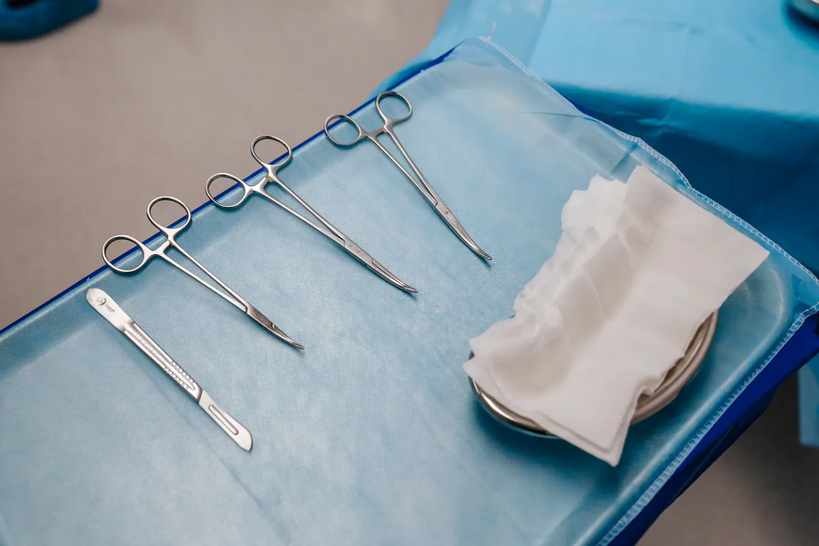 A scalpel and three pairs of surgical forceps laid out on a blue sterile drape beside folded gauze, the basic instruments a plastic surgeon trainee learns first