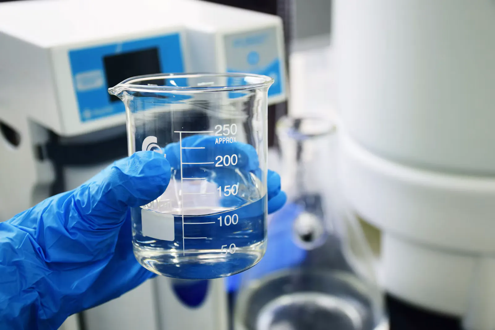 Hand in blauem Einweghandschuh hält ein Becherglas mit klarer Flüssigkeit vor Laborgeräten, Symbolbild für die Titerkontrolle nach der Hepatitis-B-Impfung