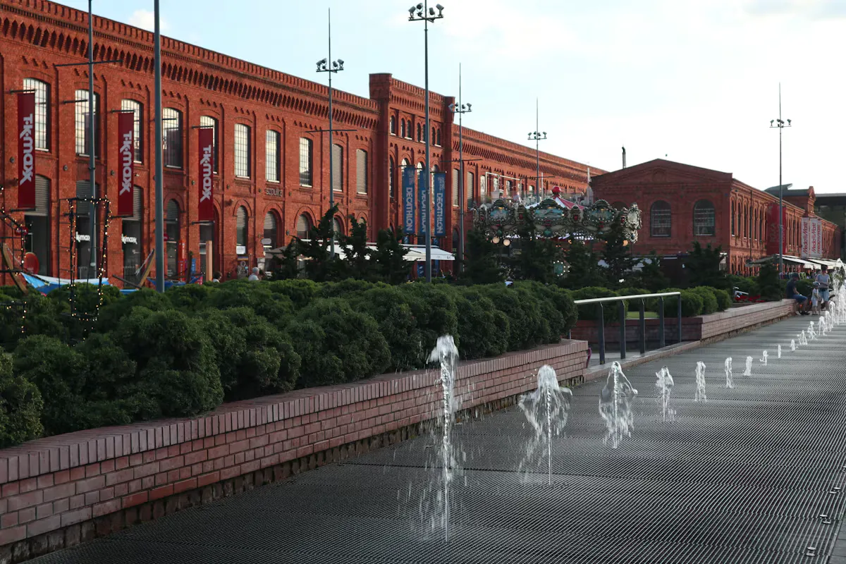 Red brick former factory buildings at Manufaktura in Lodz, with a row of small water fountains and green hedges in the foreground