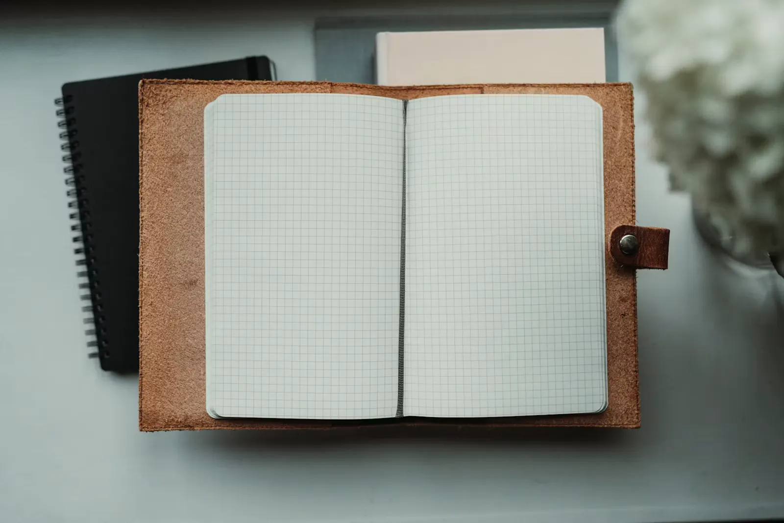 An open leather bound notebook showing blank grid ruled pages, resting on a pale desk beside a black spiral notebook