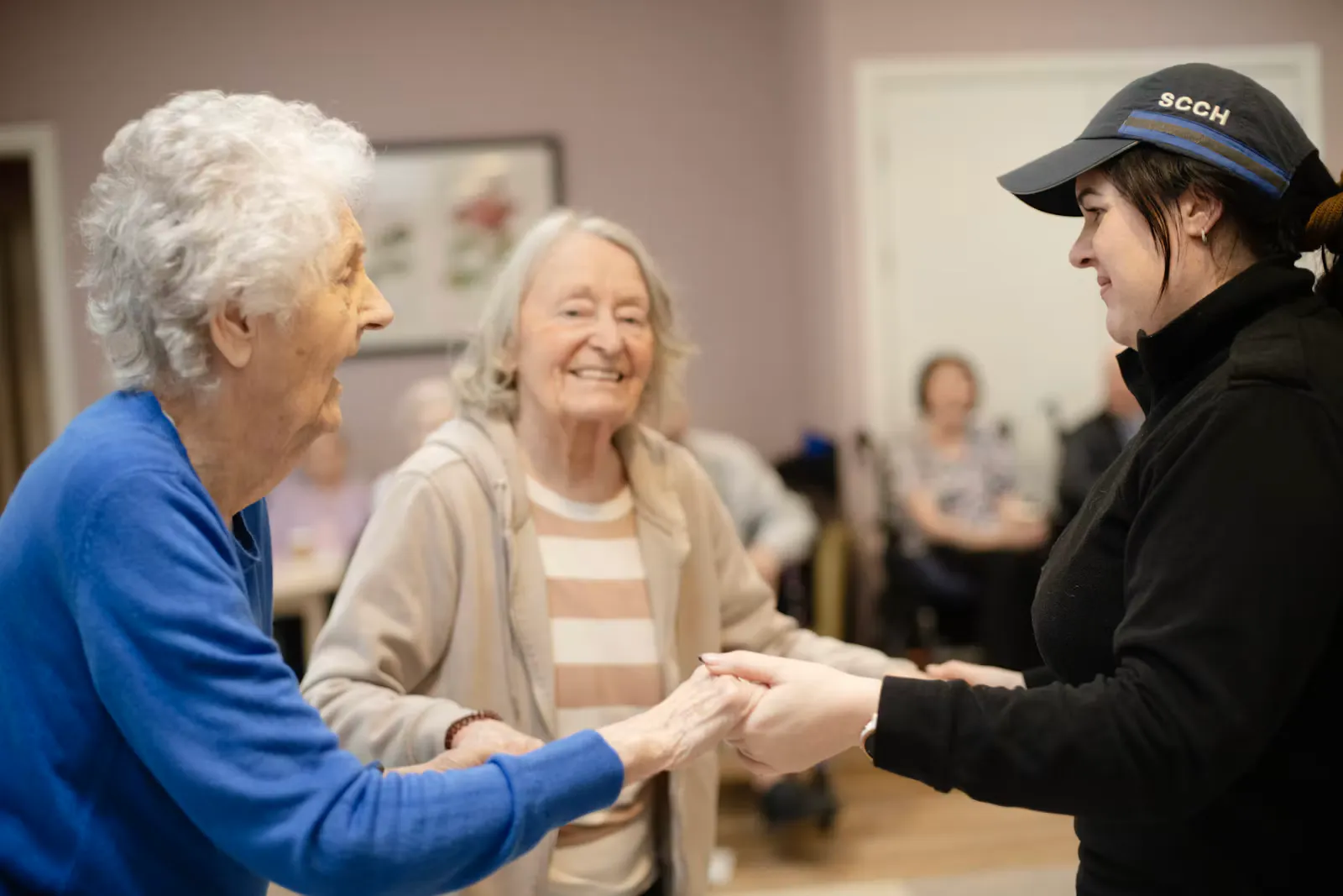A young woman in a black jacket and cap holds hands with two smiling older women in a care home common room