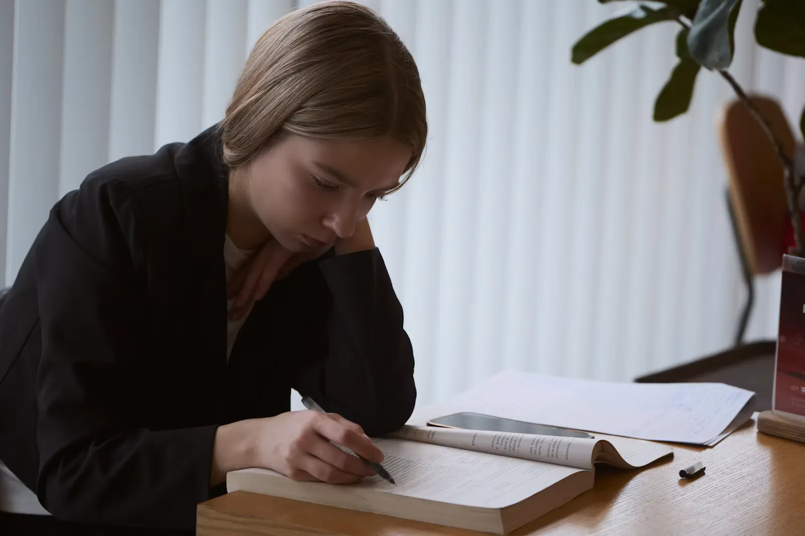 Junge Frau im dunklen Blazer lernt konzentriert mit Stift in einem aufgeschlagenen Buch an einem Holztisch