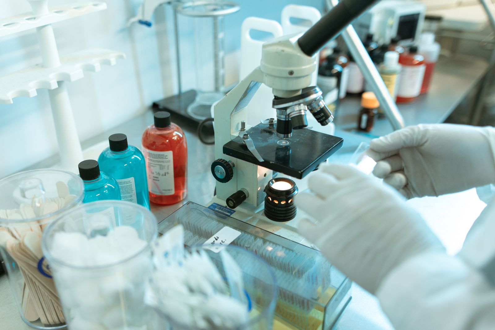 A researcher in a white coat looking into a microscope beside reagent bottles and lab equipment