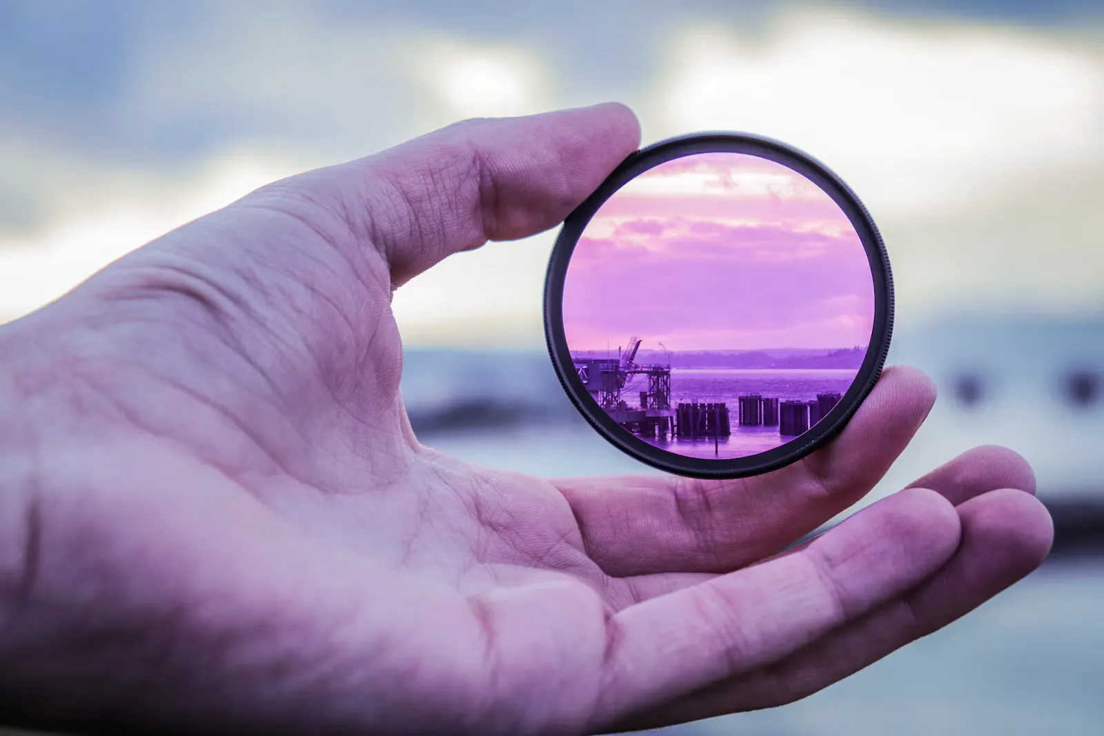 Round glass filter held up in an open hand, with a harbour and sky seen through it in purple, a reminder that ray optics is about where light goes through glass
