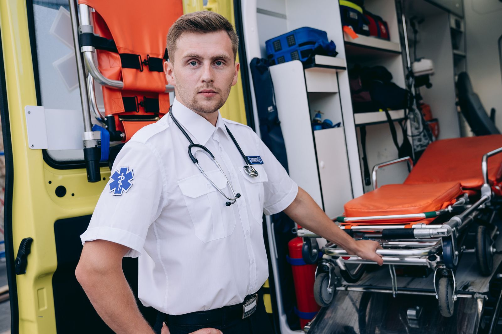 Rettungssanitäter mit Stethoskop steht am offenen Heck eines Rettungswagens neben einer Trage
