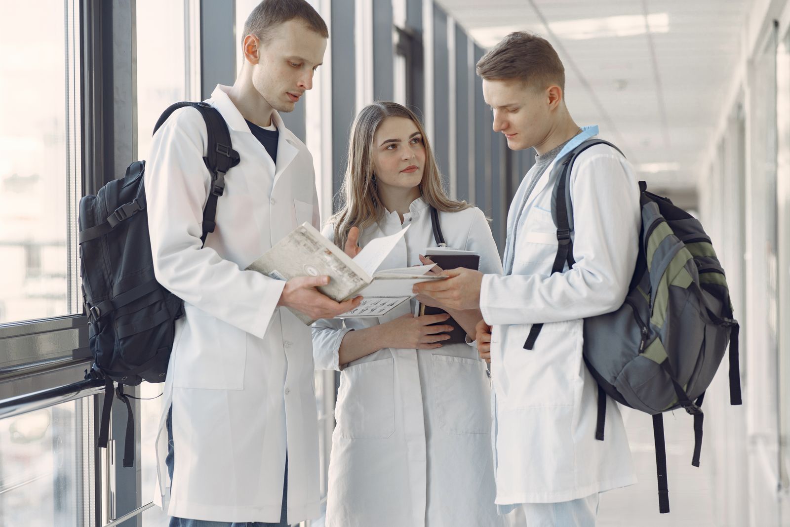 Three medical students in white coats with backpacks comparing notes and an open book in a bright hospital corridor