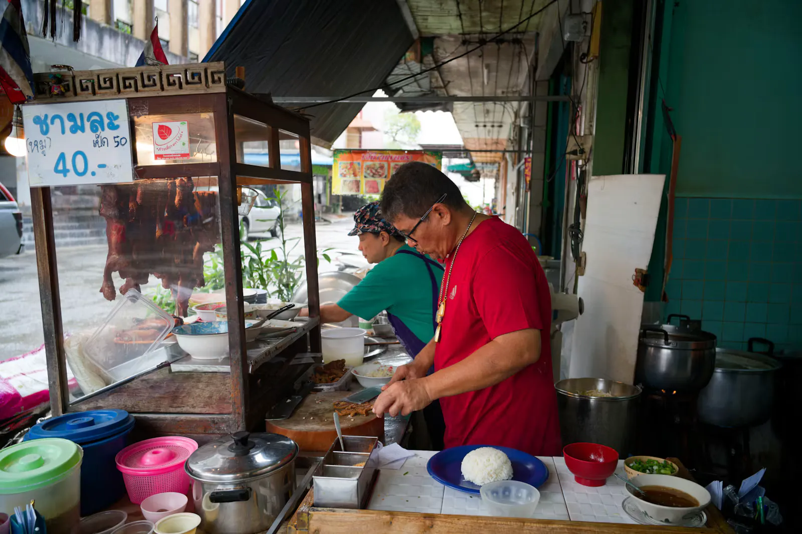Two cooks preparing rice and meat at an open street food stall with pots, bowls and a glass display case