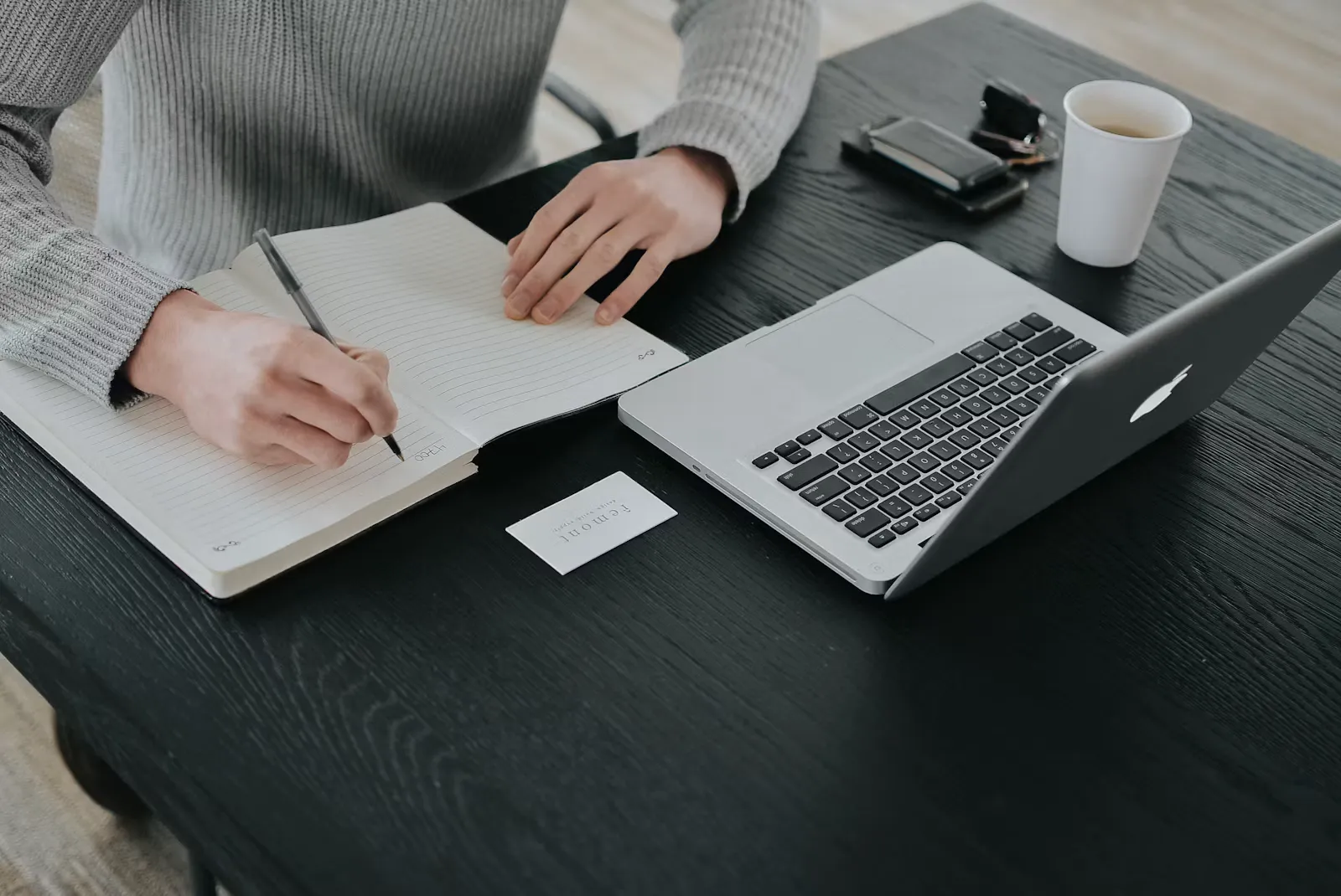 A person in a grey sweater writing notes at a dark desk, checking details before using the NEET correction window