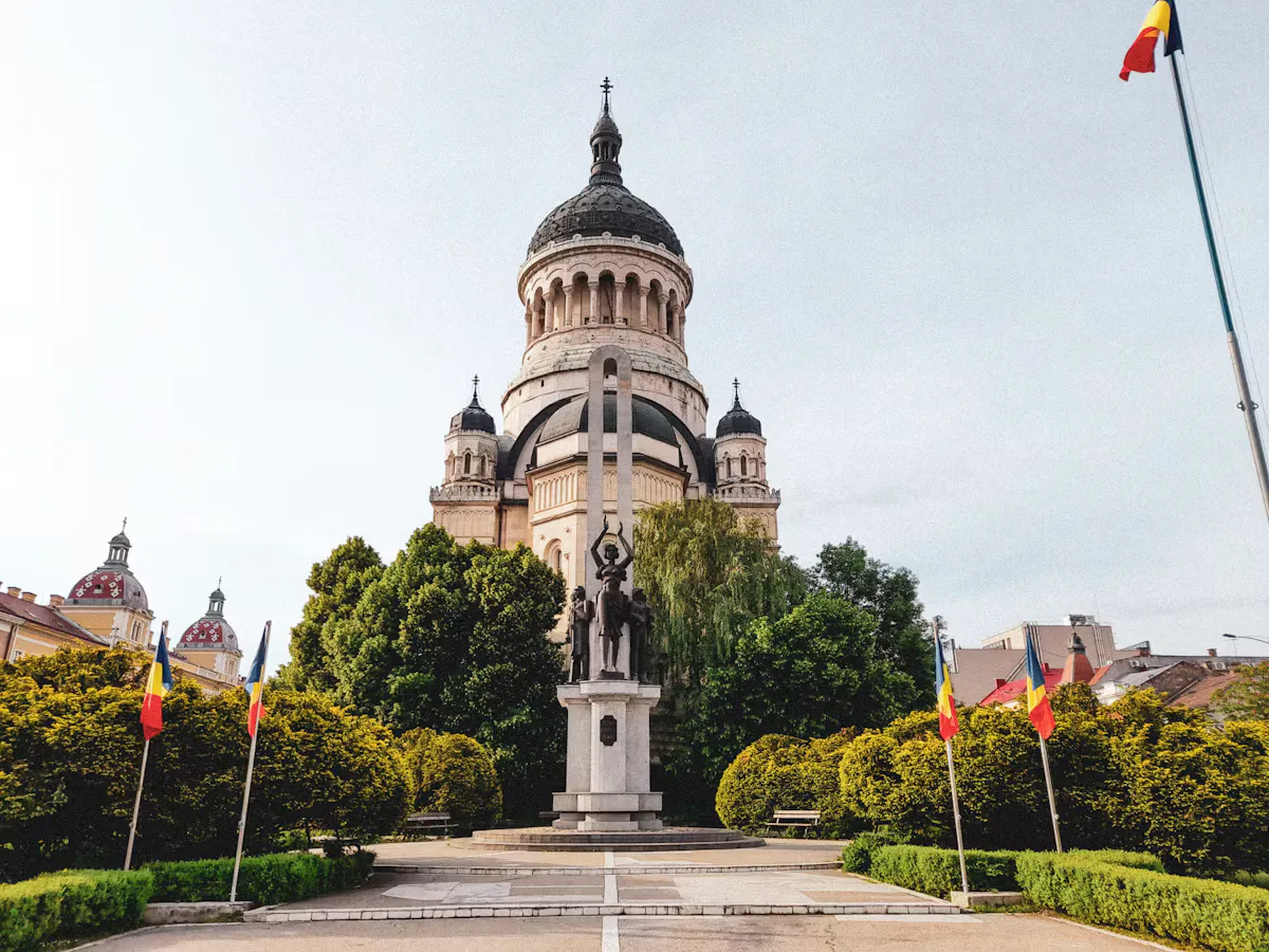 Orthodox Cathedral of Cluj-Napoca with its large dome behind the Avram Iancu monument, framed by trimmed hedges and Romanian flags on a sunny day