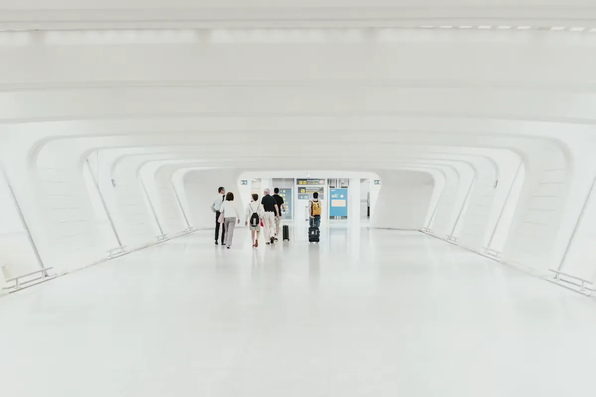 A small group of travellers with backpacks and a rolling suitcase walking through a long, bright white arched corridor towards departure screens