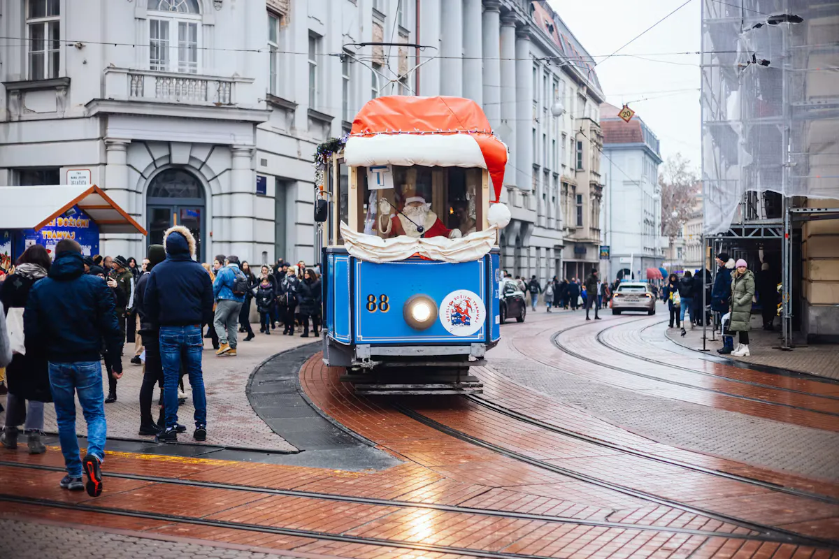 A blue tram decorated with a red and white Santa hat rolling through the centre of Zagreb in winter, with people in coats walking past old city buildings