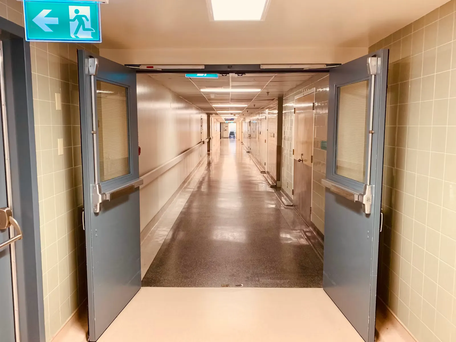 An empty hospital corridor with open double doors and a green exit sign overhead