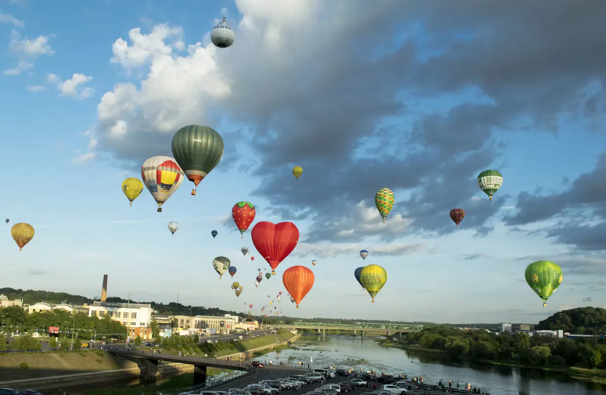 Dozens of colourful hot air balloons, including a red heart shaped one, rising over the river in Kaunas, Lithuania, on a summer evening with a bridge and green banks below