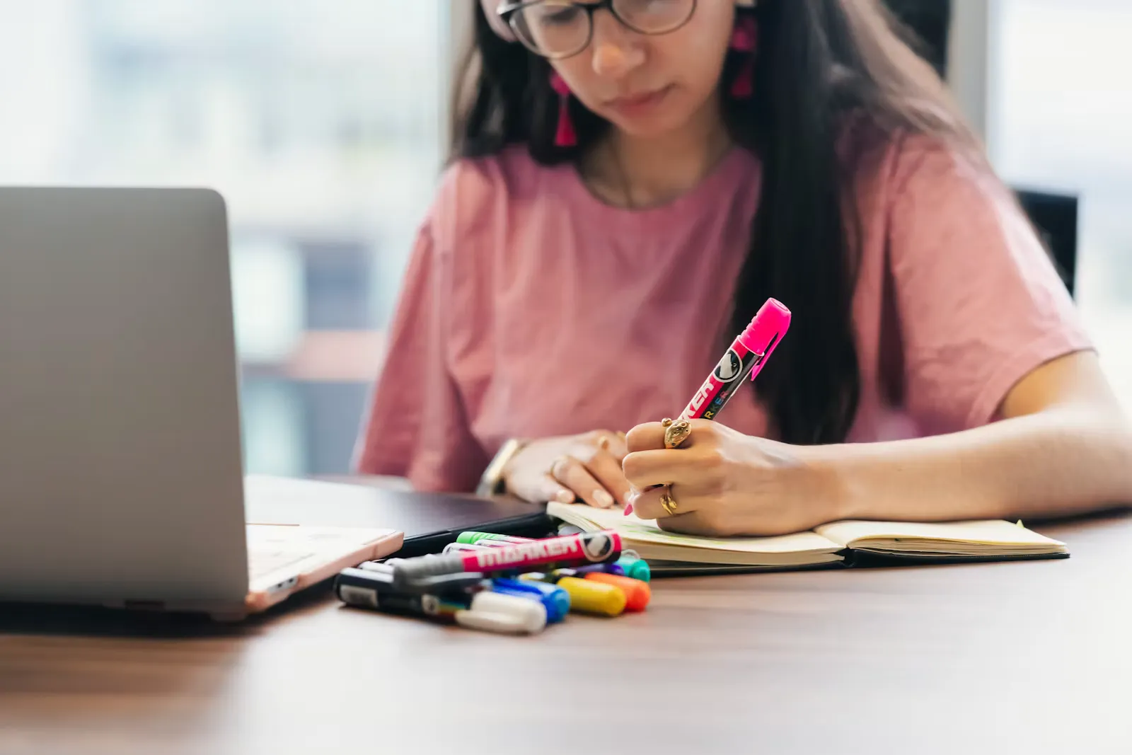 Student writing in a notebook with a pink highlighter, with coloured pens and a laptop on the desk