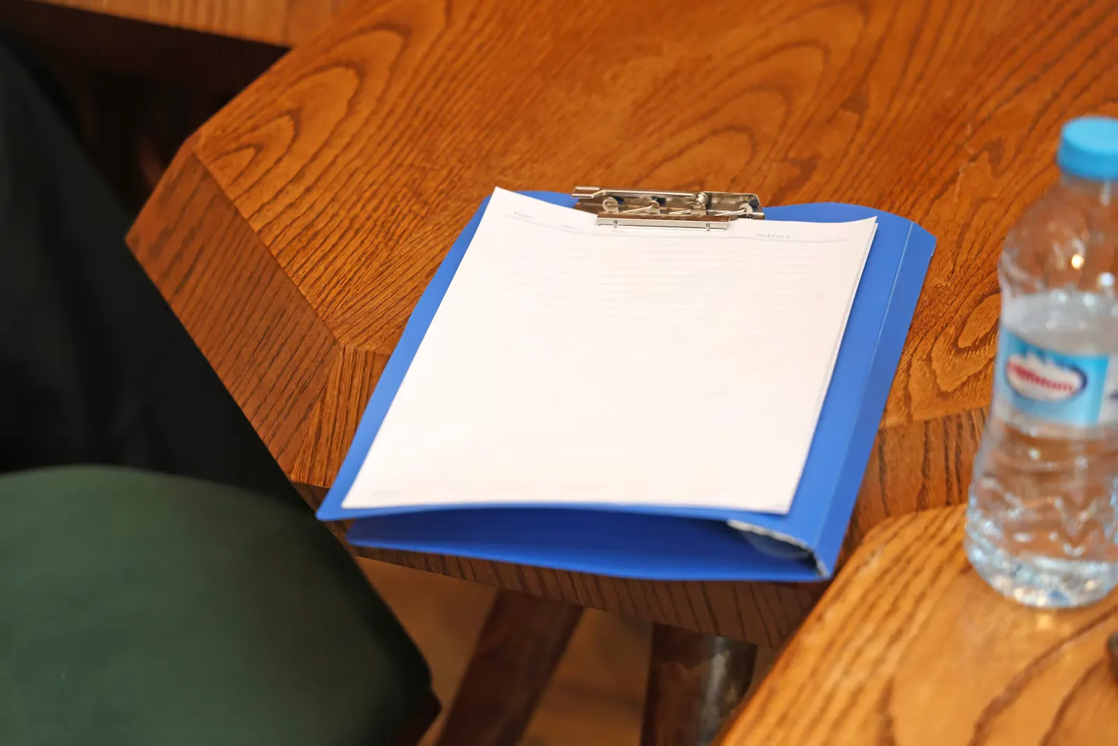A blue clipboard folder holding blank lined paper on a wooden table, with a water bottle beside it