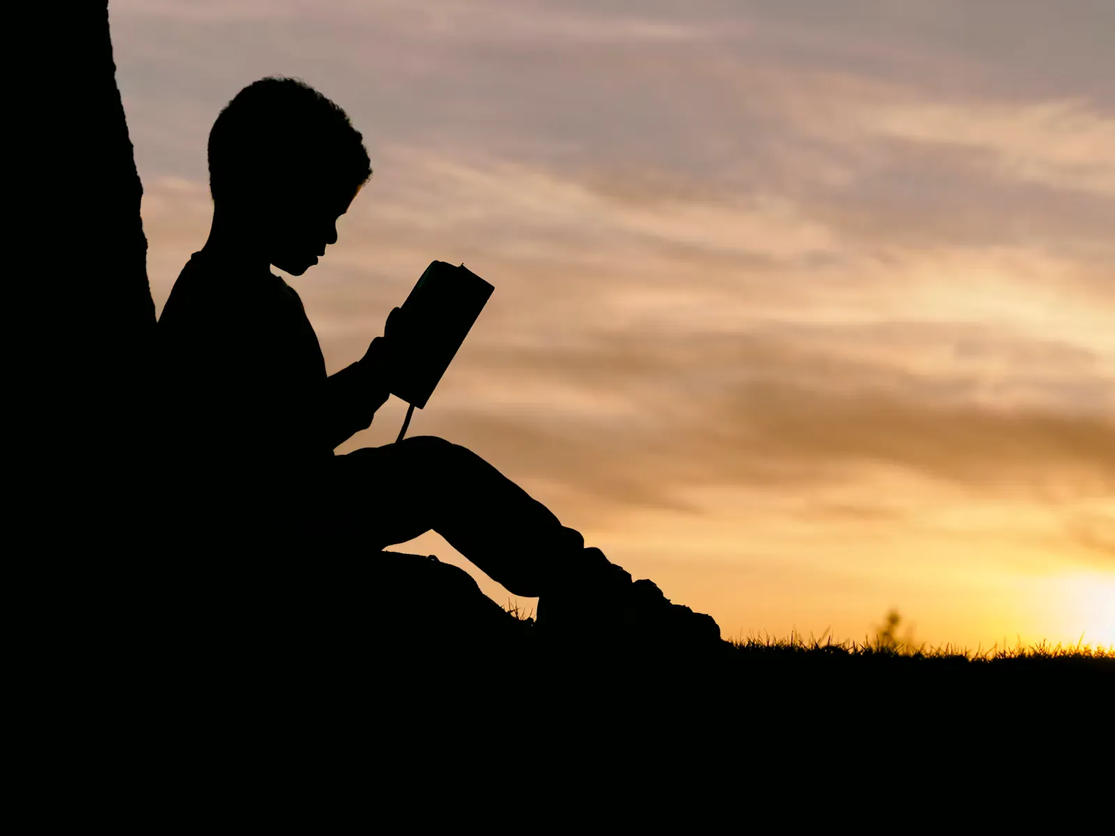 A child reading a book outdoors at sunset, silhouetted against the sky