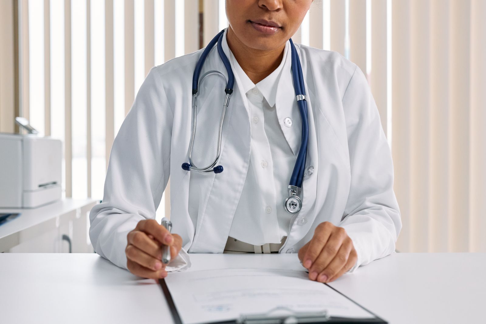 A doctor with a stethoscope writing notes at a desk in a clinic office