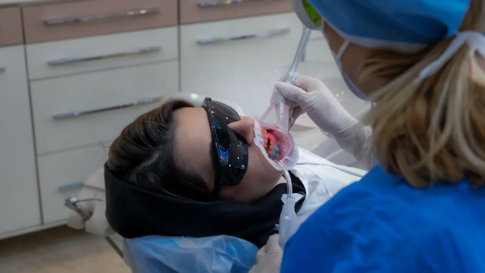 Zahnärztin mit Haube und Mundschutz behandelt eine Patientin mit Schutzbrille und Absaugung im Behandlungszimmer