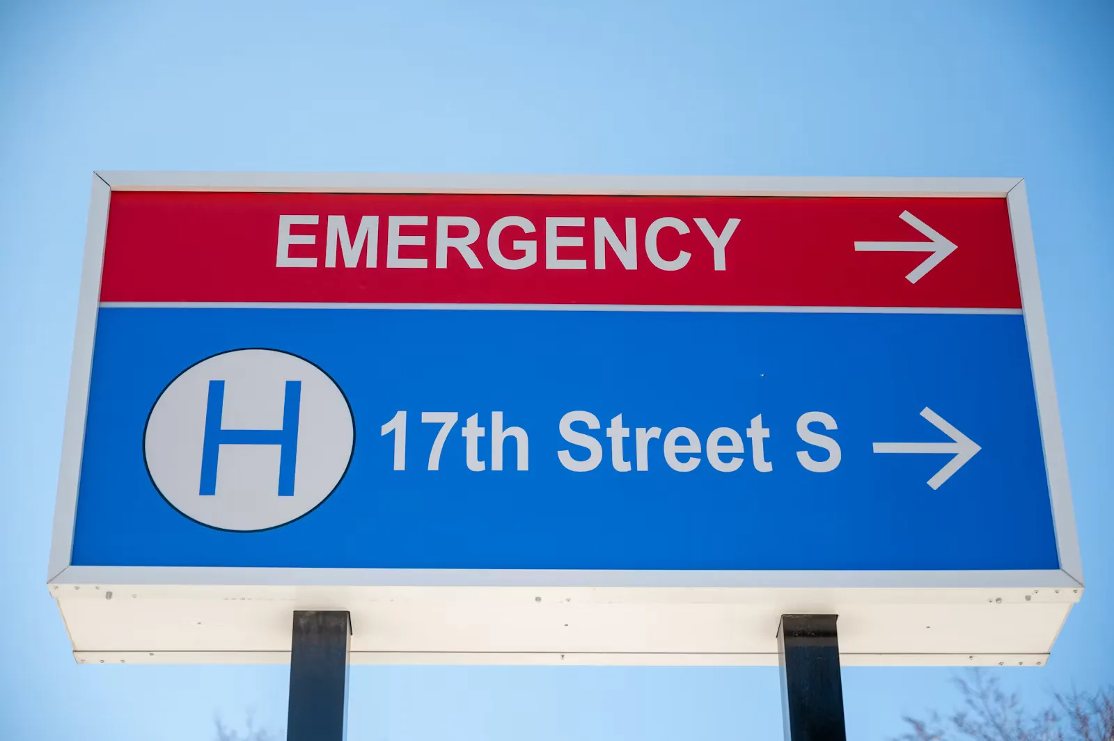 A red and blue roadside hospital sign reading EMERGENCY with an arrow, above a white H symbol and a street name, against a clear blue sky