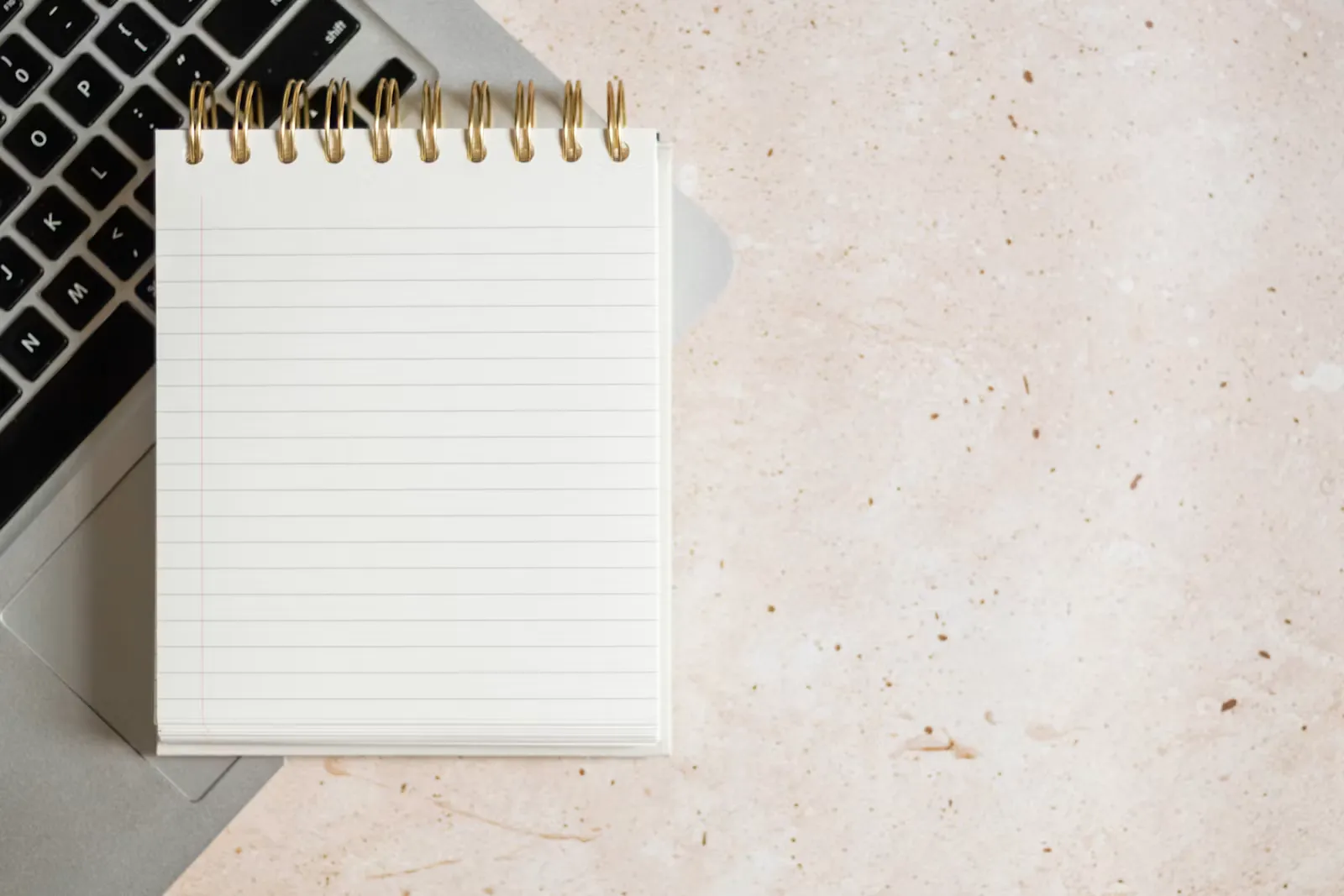 A spiral notepad with blank lined pages lying on a silver laptop keyboard on a pale speckled desk
